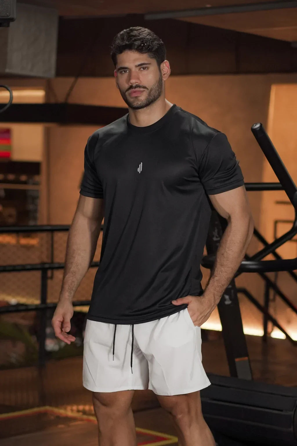 Homem usando camiseta esportiva preta e shorts branco em academia, moda fitness masculina para treino.