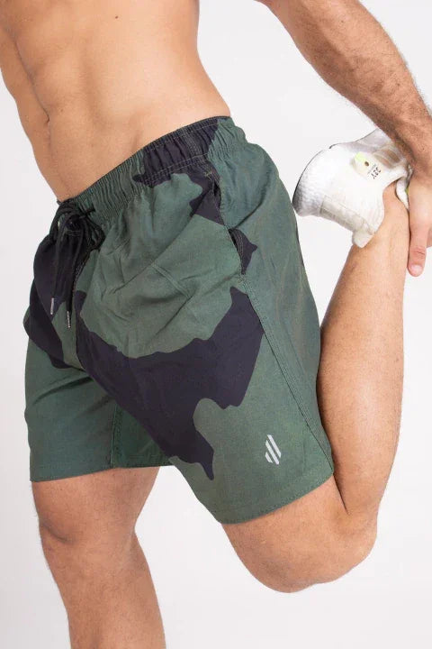 Homem usando bermuda esportiva verde com estampa camuflada e tênis branco alongando a perna.